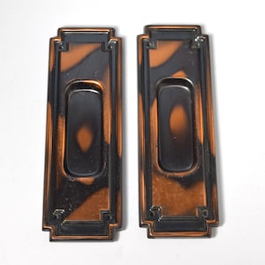 May include: Two antique door pulls with a black and copper finish. The pulls have a rectangular shape with a rounded opening in the center.