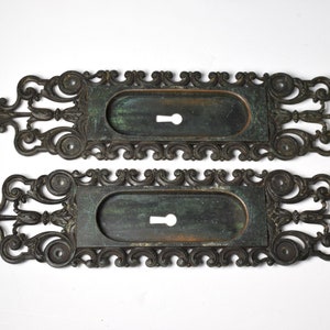 May include: Two antique ornate metal door plates with keyhole cutouts. The plates are a dark bronze color with a green patina.