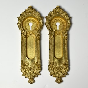 May include: Two ornate gold-toned keyhole covers with intricate floral designs. The covers are rectangular with a keyhole in the center and a decorative top.