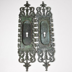 May include: Two antique ornate metal door plates with keyhole openings. The plates are a dark green color with intricate scrollwork and a decorative design at the top.