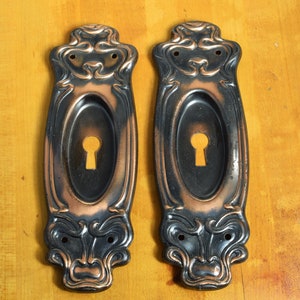 May include: Two antique keyhole covers with an ornate design. The covers are made of metal and have a black and copper finish.