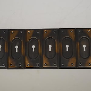 May include: A row of eight antique bronze-toned keyhole covers. Each rectangular cover has an oval keyhole with a white key outline. The edges have a gold-colored trim, and each has four screw holes.