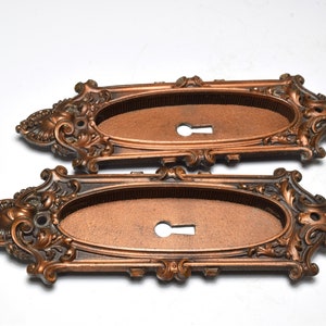 May include: Two ornate, bronze-colored keyhole covers with an oval center and decorative scrollwork. The covers have a keyhole and are likely for antique furniture or doors.