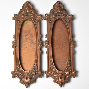 May include: Two antique oval-shaped door plate covers with ornate, detailed, and intricate designs. The plates are made of a copper-toned metal and have a worn, aged look.
