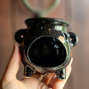 Op de afbeelding: Een zwarte keramische ketel met drie poten, twee handvatten aan de zijkant en een grote opening. De ketel heeft oranje en witte spikkels. De ketel wordt in de hand van een persoon gehouden.