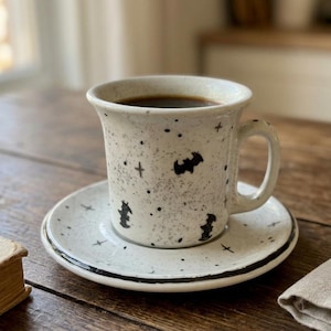 Op de afbeelding: Een gespikkelde, gebroken witte keramische koffiekop met bijpassende schotel, voorzien van een handgeschilderd patroon van kleine zwarte vleermuizen, sterren en stippen. De kop heeft een gebogen oor en de schotel heeft een dunne zwarte rand.