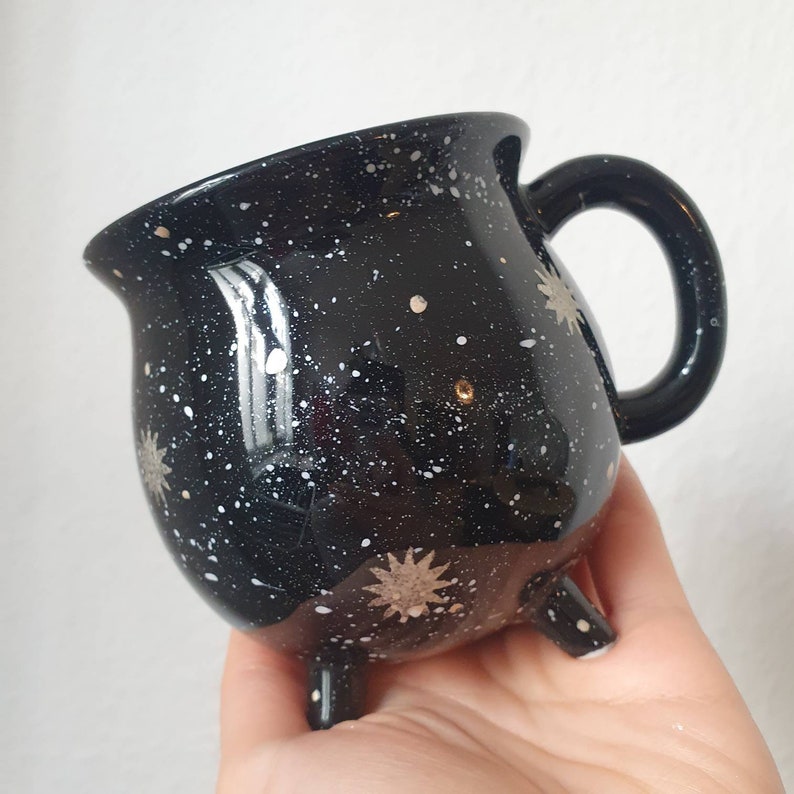 May include: Black ceramic mug shaped like a cauldron with white and silver stars and speckles. The mug has three small feet.