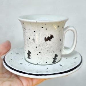 Pode incluir: Conjunto de chávena e pires de cerâmica branca com um padrão salpicado e desenhos de morcegos e cruzes pretas. A chávena tem uma pega e o pires tem uma borda preta. Um design assustador com tema de Halloween.