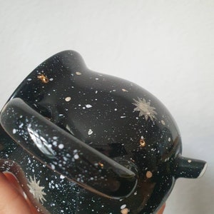 May include: Black ceramic teapot with a white and gold speckled design. The teapot has a handle and a spout.