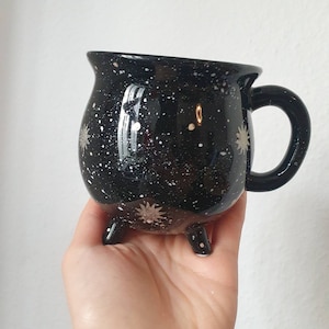 May include: Black ceramic mug with a white speckled galaxy design and a handle. The mug has three small feet.
