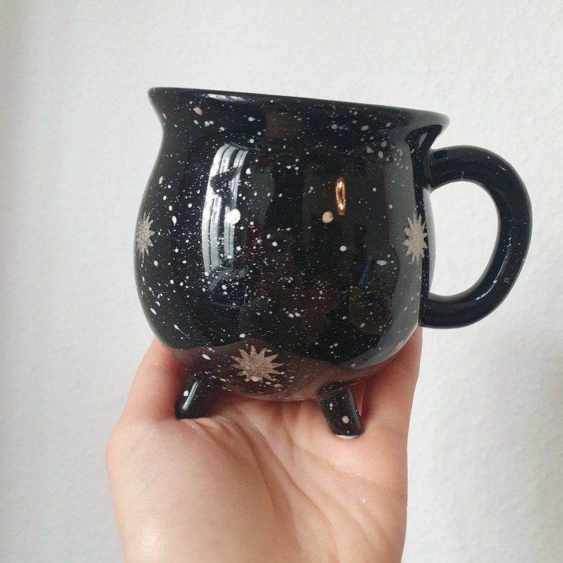 May include: Black ceramic mug with a white speckled galaxy design and a handle. The mug has three small feet.
