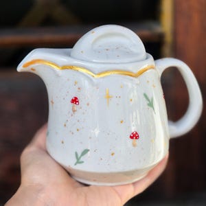 Könnte beinhalten: Eine weiße Keramik-Teekanne mit Deckel, Griff und Ausguss. Die Teekanne ist mit roten Pilz- und grünen Blattmotiven, goldenen Akzenten und goldenen Sprenkeln verziert. Die Teekanne wird in einer Hand gehalten.