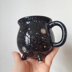 May include: Black ceramic mug shaped like a cauldron with white and gold star accents. The mug has three legs and a handle.
