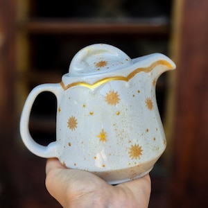 Könnte beinhalten: Eine Keramikkrug mit Deckel, Griff und Ausguss. Der Krug ist cremefarben mit goldenen Sternen und Punktmustern. Der Rand und der Ausguss sind goldfarben verziert. Der Krug wird in einer Hand gehalten.