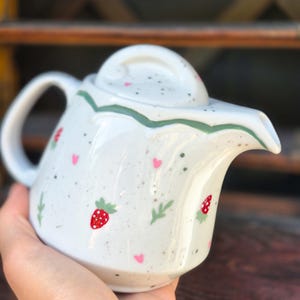 Könnte beinhalten: Weiße Keramik-Teekanne mit Deckel, Henkel und Ausguss. Die Teekanne ist mit roten Erdbeeren, rosa Herzen und grünen Blättern verziert. Der Rand der Teekanne ist mit einer grünen Wellenlinie umrandet. Die Teekanne wird in einer Hand gehalten.