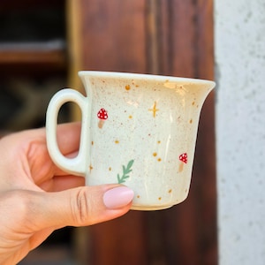Peut inclure: Mug en céramique crème avec une finition mouchetée, orné de motifs de champignons rouges et de feuilles vertes peints à la main. La tasse a une anse incurvée et un bord évasé. La tasse est tenue dans une main.