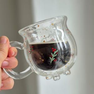 Könnte beinhalten: Eine klare Glas Tasse mit Henkel, verziert mit goldenen Punkten und gemalten Stechpalmenzweigen. Die Tasse ist mit dunklem Kaffee gefüllt.