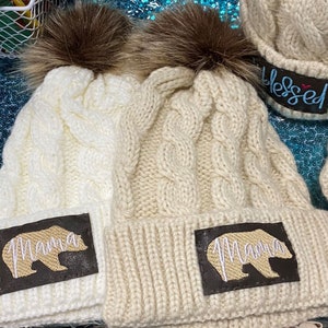 May include: Two white and beige knit winter hats with faux fur pom poms. The hats have a cable knit pattern and a brown patch with the word "Mama" and a bear silhouette embroidered on them.