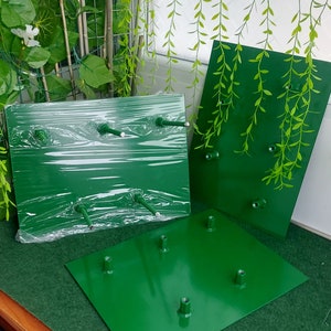 May include: Several bright green metal display bases with attached posts are shown. One is wrapped in clear plastic. The bases are rectangular and square, and appear to be for displaying items. The background includes artificial greenery.