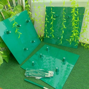 May include: Three green metal square plates with multiple metal posts, likely for a garden structure. A bundle of metal rods and a single rod are also present. The plates are a glossy green color, and the background includes greenery.