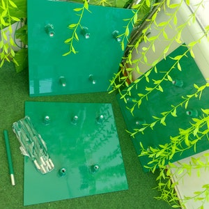 May include: Three square, bright green metal plates with six bolt holes each. A bundle of bolts and a green rod are next to one of the plates. The plates are arranged on a green surface with green leafy vines in the background.