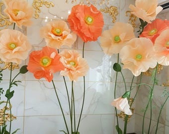 Könnte beinhalten: Eine Sammlung großer, freistehender künstlicher Blumen in Pfirsich- und leuchtenden Orangetönen. Jede Blume hat geschichtete, gerüschte Blütenblätter, einen grünen Staubfaden in der Mitte und einen dünnen grünen Stiel auf einem runden Sockel.