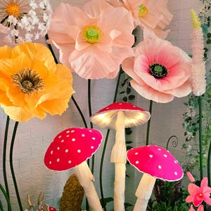 Puede incluir: Una colección de grandes flores decorativas de papel en tonos melocotón, rosa y amarillo, junto a tres esculturas de setas iluminadas con sombreros rojos brillantes y puntos blancos, y una pequeña figura de ciervo texturizada sobre base verde.
