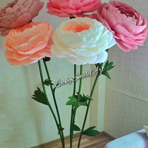 May include: A collection of large, paper peony flowers in shades of pink, peach, and white. The flowers are mounted on green stems with leaves, and the base is a square of green material. The text "Lucky Smile Art" is visible on the stems.