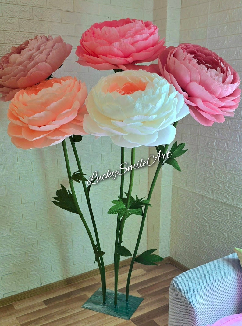 May include: A collection of large, paper peony flowers in shades of pink, peach, and white. The flowers are mounted on green stems with leaves, and the base is a square of green material. The text "Lucky Smile Art" is visible on the stems.