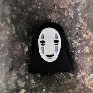 以下が含まれることがあります： 千と千尋の神隠し のカオナシの刺繍パッチ。黒と白で刺繍されています。パッチは紫色の背景の上にあります。