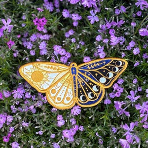 May include: Embroidered butterfly patch with a celestial design. One wing features a sun and white details, the other wing has a moon phase design with stars on a navy blue background. The patch has a gold border and is set against a background of purple flowers.