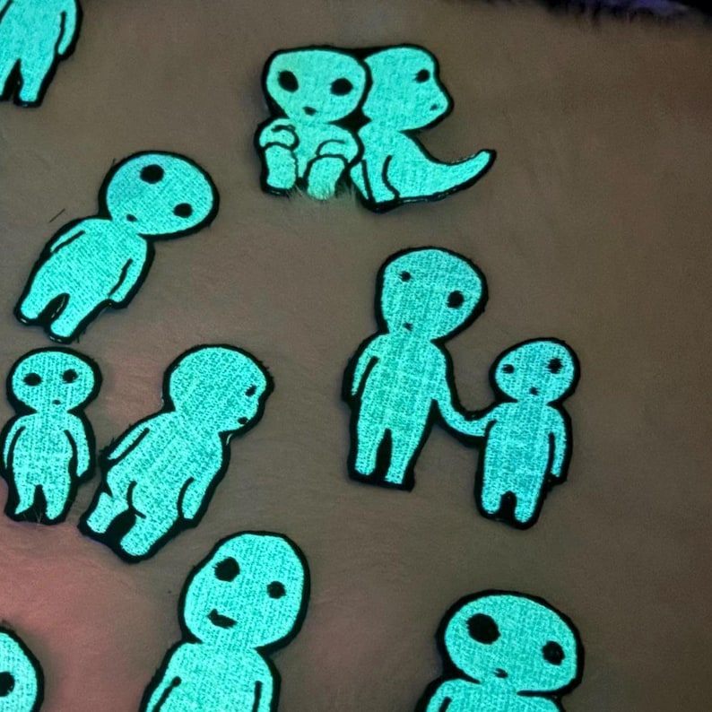 May include: A collection of glow-in-the-dark embroidered patches featuring small, humanoid figures with large heads and black eyes. The patches are light green and have black stitched outlines, depicting individual figures and pairs.