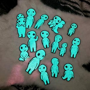 May include: A collection of glow-in-the-dark embroidered patches featuring various small, stylized humanoid figures with large round eyes and simple bodies. The patches emit a bright cyan light against a dark background, showcasing diverse poses.