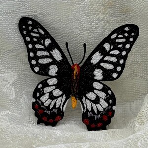 May include: An embroidered butterfly patch featuring a black base with white wing patterns, a central body with yellow and red accents, and small red spots along the lower edges of the wings. The design is symmetrical with two thin antennae.
