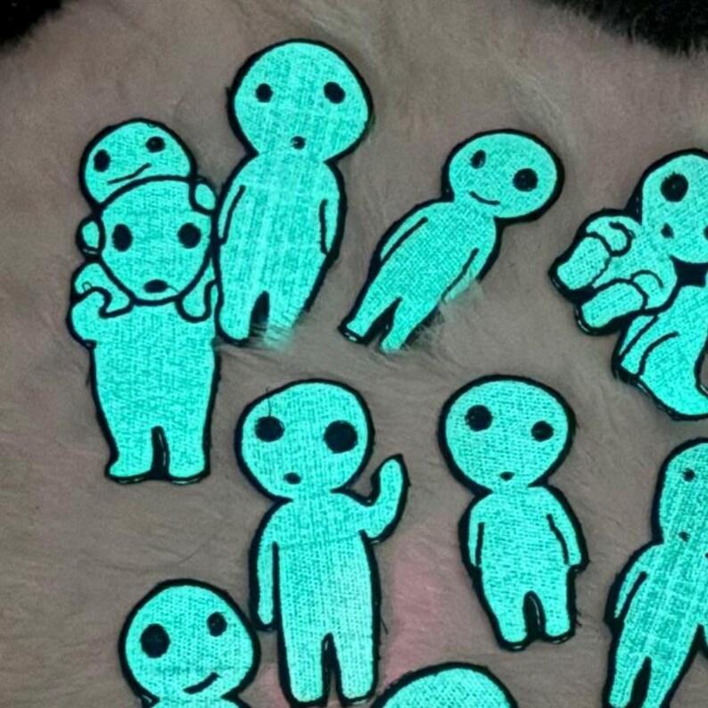 May include: A collection of glow-in-the-dark embroidered patches featuring stylized, light-blue figures with round heads and simple facial features. The patches depict various poses, including standing figures and one figure carrying another on its back.
