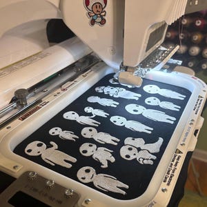 May include: A white embroidery machine holds a black fabric hoop featuring multiple white stitched figures resembling forest spirits. The machine has a small Toad character sticker on the top and a metal frame securing the fabric in place.