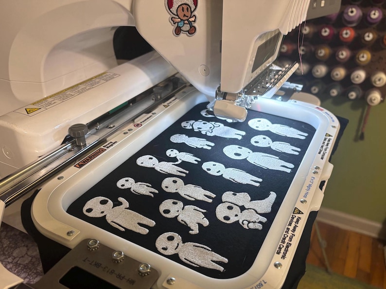 May include: A white embroidery machine holds a black fabric hoop featuring multiple white stitched figures resembling forest spirits. The machine has a small Toad character sticker on the top and a metal frame securing the fabric in place.