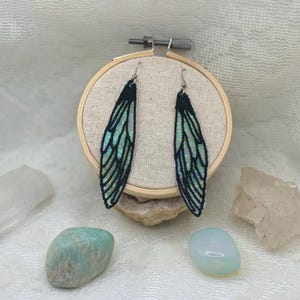Può includere: Un paio di orecchini ad ali iridescenti fatti a mano. Gli orecchini presentano un contorno nero con una sfumatura di colore cangiante, verde e turchese. Le ali sono sospese a ganci color argento.