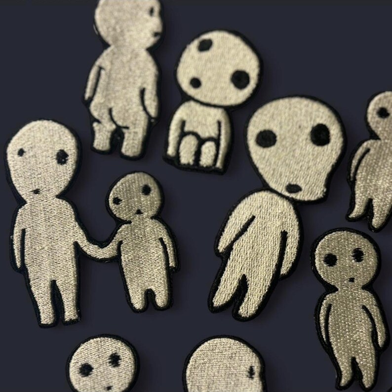 May include: A collection of embroidered patches featuring stylized, pale beige humanoid figures with simple black circular eyes and mouths. The figures vary in posture, with some standing alone, some holding hands, and others shown as partial heads.