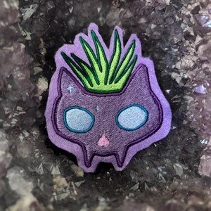May include: Purple embroidered cat with green plant growing out of its head. The cat has sparkly eyes and a pink nose.