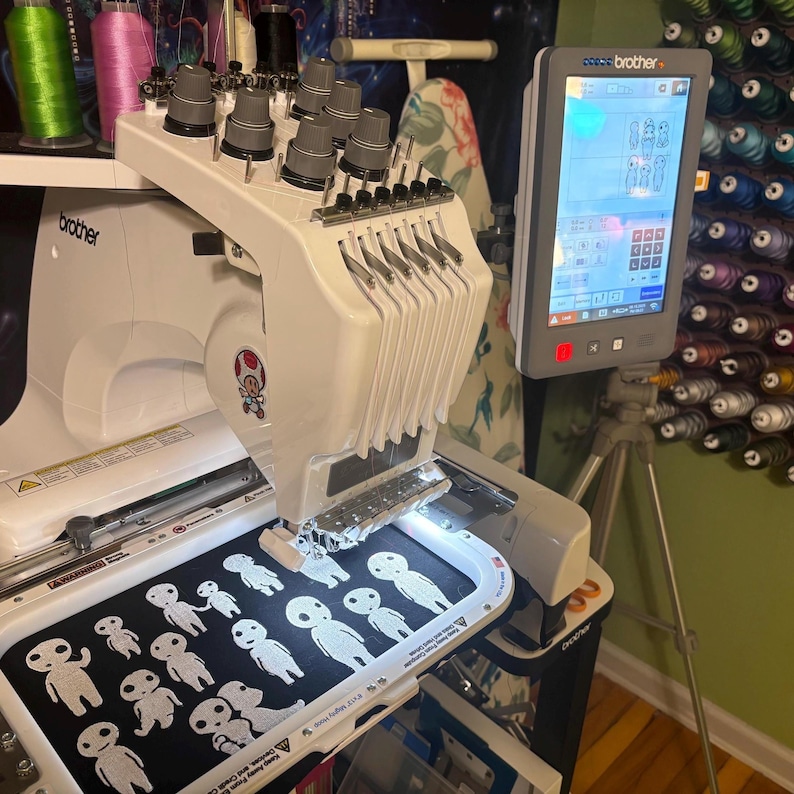 May include: A white Brother multi-needle embroidery machine with a digital touchscreen display. The machine is actively stitching white line-art figures of small, round-headed characters onto a black fabric hoop, showing multiple repeating designs.