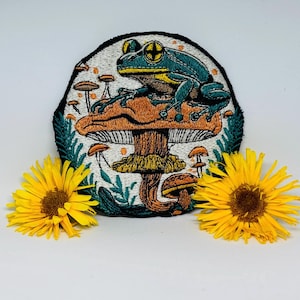 May include: Embroidered patch featuring a blue and yellow frog perched on an orange mushroom. The patch is surrounded by other mushrooms and green foliage, with a black border. Two yellow flowers are placed on either side of the patch.