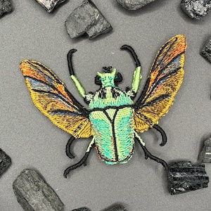 Puede incluir: Parche bordado de un escarabajo con las alas extendidas. El escarabajo tiene un cuerpo verde y turquesa con patas y antenas negras. Las alas son amarillas, naranjas y azules. El parche está rodeado de piedras grises y negras.