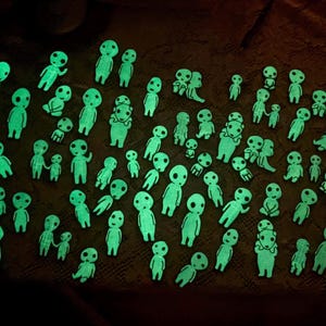 May include: A large collection of small, glow-in-the-dark figurines shaped like stylized forest spirits. Each figure features a smooth, rounded head with large, hollow eyes and a simple body, emitting a bright, neon green light in the darkness.