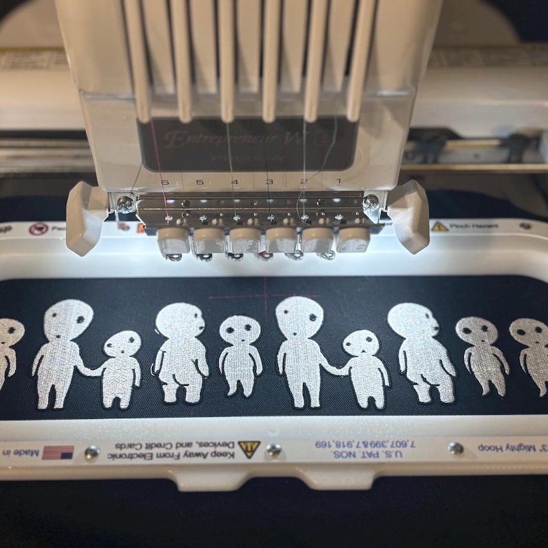 May include: A white embroidery machine hoop holds black fabric featuring a repeating white stitched pattern of small, stylized humanoid figures. The machine head is positioned above the hoop, showing needles and thread guides in a white housing.