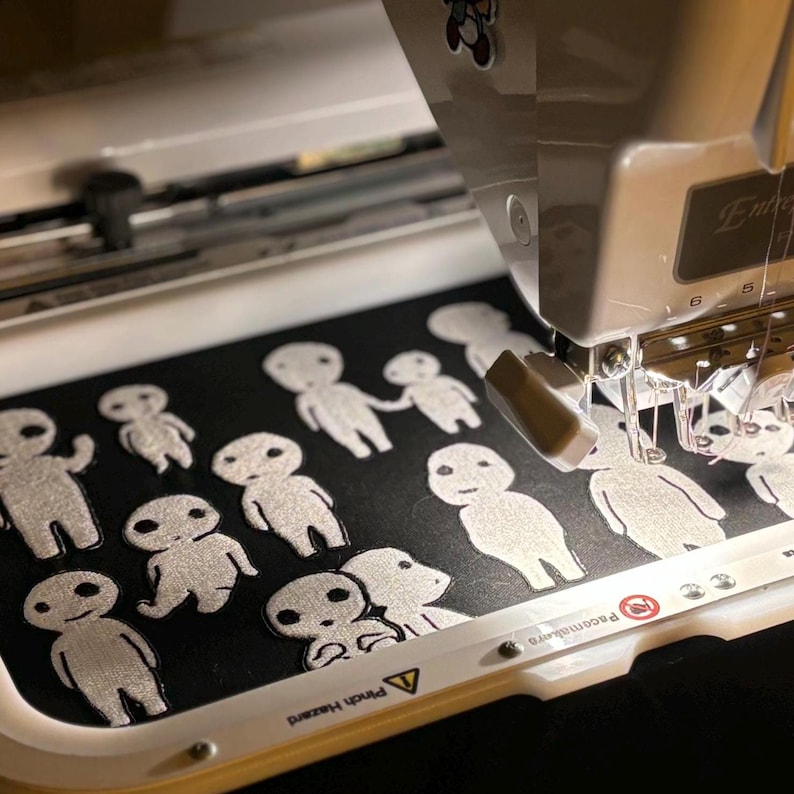 May include: An embroidery machine stitching multiple white, stylized humanoid figures onto a black fabric surface held within a rectangular hoop. The machine features a white casing with visible mechanical components and a warning label.