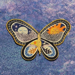 May include: Embroidered butterfly patch with a celestial design. The wings feature a sun, moon, stars, and ocean waves in shades of blue, orange, and yellow. The patch has a black border with gold trim.