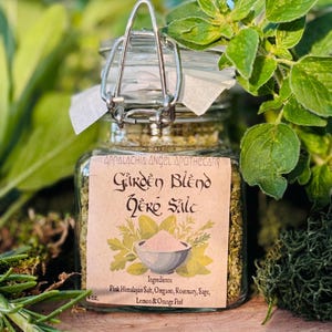 May include: A clear glass jar with a metal clasp, filled with green herbs and pink salt. The label reads "Garden Blend Herb Salt" with a list of ingredients: Pink Himalayan Salt, Oregano, Rosemary, Sage, Lemon & Orange Peel. The jar is surrounded by green foliage.
