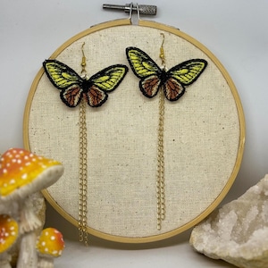 Puede incluir: Un par de pendientes colgantes con motivos de mariposas bordadas en tonos amarillo, naranja y negro. Cada mariposa está unida a un gancho de metal dorado y presenta una cadena dorada larga y delicada que cuelga del borde inferior.