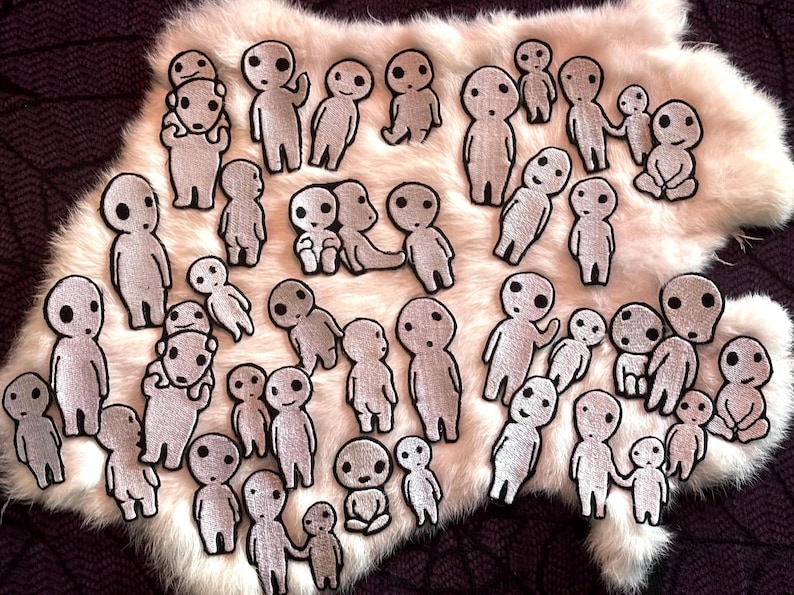 May include: A collection of gray embroidered patches featuring stylized figures with large, round eyes. The patches are in various poses, some standing, some holding hands, and some sitting. They are arranged on a white, fluffy surface.